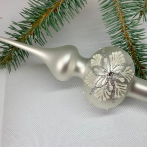 May include: A silver Christmas tree topper with a snowflake design. The glass ornament has a long, pointed tip and a round base with a glittery finish. A sprig of evergreen is in the background.