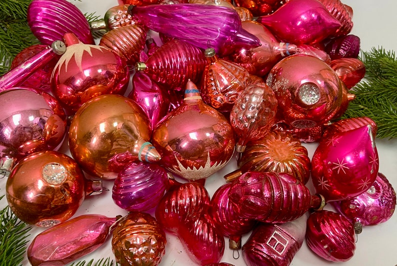 May include: A collection of vintage pink and red glass Christmas ornaments. The ornaments are all different shapes and sizes, and some have a sparkly finish.