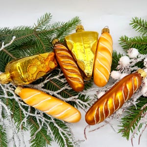 May include: A collection of glass Christmas ornaments in the shape of food items. The ornaments include a cheese wedge, a bottle, and several baguettes. The ornaments are gold, orange, and white, and are arranged on a bed of greenery.