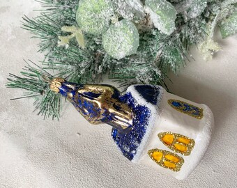 May include: A glass Christmas ornament shaped like a house with a blue and gold roof and white walls. The windows are outlined in gold glitter. The ornament is decorated with artificial snow and rests near a frosted evergreen branch.