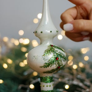 May include: White glass Christmas tree topper with green glitter pine branches and red berries. The topper has a white base with green glitter stripes.