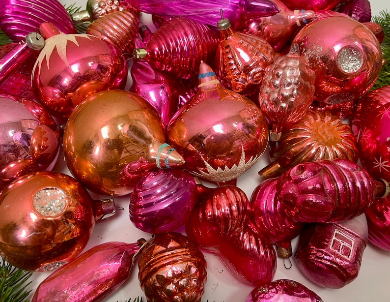 May include: A collection of vintage pink and gold glass Christmas ornaments. The ornaments are all different shapes and sizes, and some have a shiny, reflective finish.