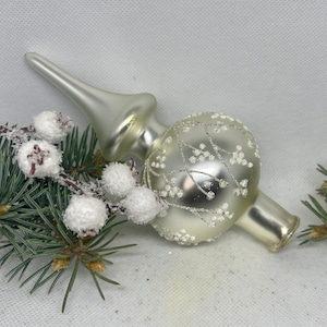 Op de afbeelding: Een witte glazen kerstboomtopper met een delicaat zilveren en wit bloemenmotief. De topper heeft een puntige bovenkant en een ronde basis.
