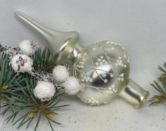 May include: A white glass Christmas tree topper with a delicate silver and white floral design. The topper has a pointed top and a round base.