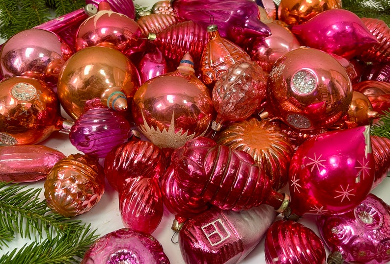 May include: A collection of vintage pink and peach glass Christmas ornaments. The ornaments are various shapes and sizes, including round, teardrop, and star shapes. Some ornaments have a shiny finish, while others have a matte finish.