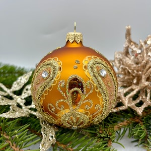 May include: A decorative orange Christmas ornament with gold glitter paisley designs and a small jewel. The ornament is resting on a bed of green pine needles and gold glitter accents. A gold cap tops the ornament.