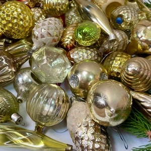 May include: A collection of vintage glass Christmas ornaments in gold and silver tones. The ornaments are various shapes and sizes, including round, teardrop, and star shapes. Some ornaments have a textured surface, while others are smooth. The ornaments are arranged on a white surface.