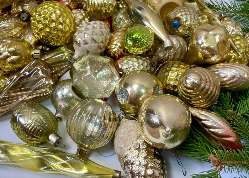 May include: A collection of vintage glass Christmas ornaments in gold and silver tones. The ornaments are various shapes and sizes, including round, teardrop, and star shapes. Some ornaments have a textured surface, while others are smooth. The ornaments are arranged on a white surface.