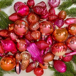 May include: A collection of vintage pink and red glass Christmas ornaments. The ornaments are in various shapes and sizes, including balls, stars, and bells. Some of the ornaments have a shiny finish, while others have a matte finish.