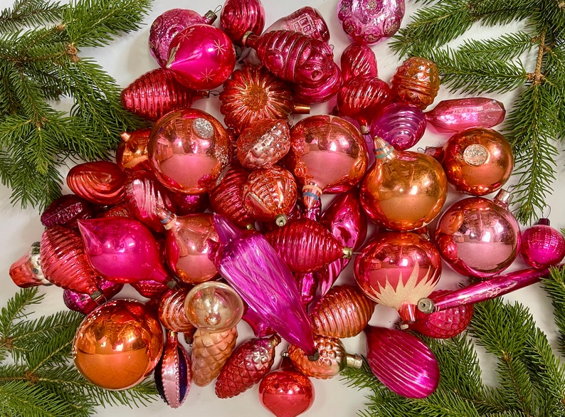 May include: A collection of vintage pink and red glass Christmas ornaments. The ornaments are in various shapes and sizes, including balls, stars, and bells. Some of the ornaments have a shiny finish, while others have a matte finish.