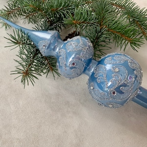 May include: A blue glass Christmas tree topper with a silver glitter design. The topper has a pointed top and a long, thin stem.