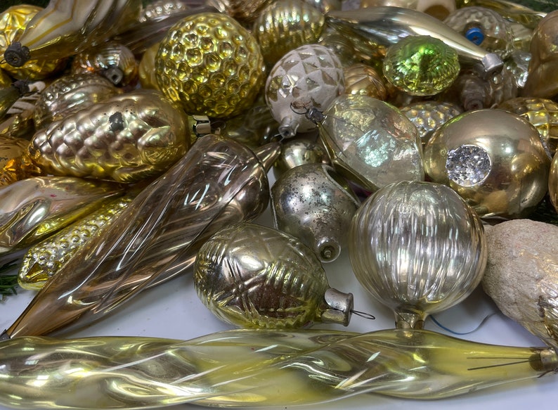 May include: A collection of vintage glass Christmas ornaments in gold, silver, and green. The ornaments are various shapes, including round, teardrop, and icicle. Some ornaments have a textured surface, while others are smooth.