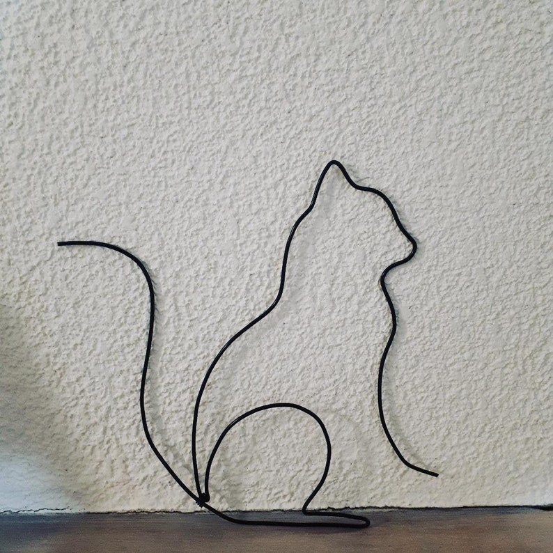 K&ouml;nnte beinhalten: Schwarze Metallskulptur einer Katze in sitzender Position. Die Katze wird durch eine durchgehende Metalllinie dargestellt. Die Skulptur ist an einer strukturierten, wei&szlig;en Wand befestigt.
