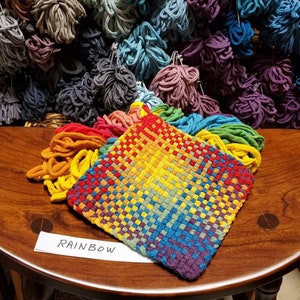 May include: A rainbow woven tapestry with a label that reads "RAINBOW". The tapestry is made of colorful yarn and is displayed on a wooden table.