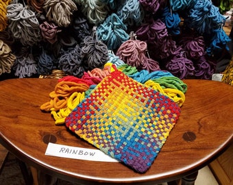May include: A rainbow woven tapestry with a label that reads "RAINBOW". The tapestry is made of colorful yarn and is displayed on a wooden table.