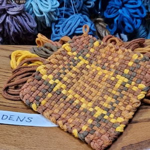 May include: A woven pot holder with a brown, yellow, and orange checkered pattern. The pot holder has fringe on all sides.