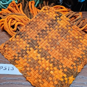 May include: A woven pot holder in shades of orange and brown, with a looped edge and a small loop for hanging. The word "COREOPSIS" is written on a white label. The pot holder is on a wooden surface.