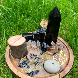 May include: A wooden incense burner with a resin-filled tray featuring dried flowers, crystals, and a black obsidian tower. The tray has a small hole for incense sticks and a gold leaf accent.