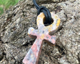 May include: A brown, yellow, and pink stone ankh pendant on a black cord necklace. The pendant is carved in the shape of an ankh, an ancient Egyptian symbol of life.