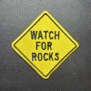 May include: Yellow diamond-shaped patch with black border and text "WATCH FOR ROCKS". The patch is embroidered with black thread on a yellow background. The patch is likely for a jacket or bag.