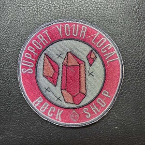 May include: Round patch with a pink border and the text "SUPPORT YOUR LOCAL ROCK SHOP". The center features a gray circle with pink crystal illustrations. The patch is on a black textured surface.