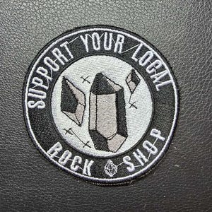 May include: Round black and white patch with the text "SUPPORT YOUR LOCAL ROCK SHOP". The patch features a gray crystal design in the center. The patch is on a black textured surface.