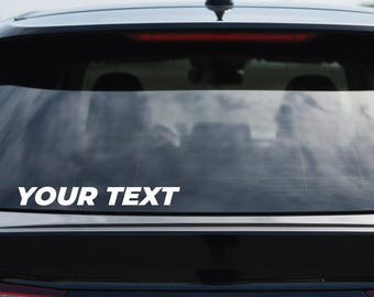 May include: A black car's rear window with the white text "YOUR TEXT" across the bottom. The car has a black antenna and red taillights. The background is a clear, blue sky.