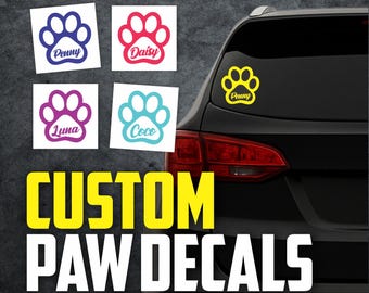 May include: Custom paw decals in various colors, including blue, pink, purple, and turquoise. Each decal features a paw print with a pet's name, such as "Penny," "Daisy," "Luna," and "Coco." The image also shows a yellow paw decal on a car.