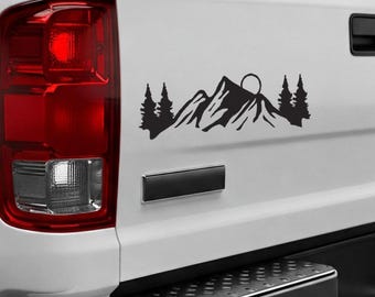 May include: Black and white silhouette decal of a mountain range with a sun rising behind it and trees on either side. The decal is applied to the back of a white pickup truck.