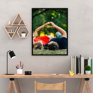 May include: A framed photograph of a couple lying on their backs in a grassy field. They are holding hands above their heads in the shape of a heart. The woman is wearing a red dress and the man is wearing a black shirt.
