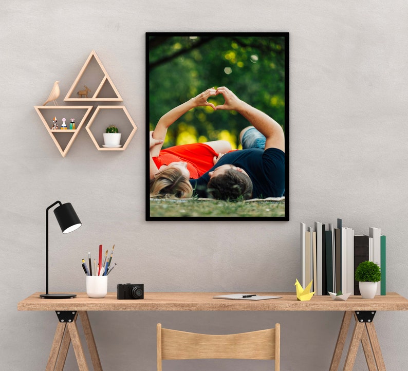 May include: A framed photograph of a couple lying on their backs in a grassy field. They are holding hands above their heads in the shape of a heart. The woman is wearing a red dress and the man is wearing a black shirt.