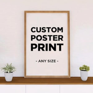 May include: A rectangular poster print in a light wood frame featuring the black text CUSTOM POSTER PRINT - ANY SIZE - on a white background. The poster is centered on a flat surface with a small green potted plant on each side.