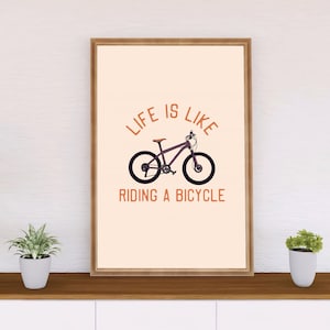 May include: A motivational poster with a beige background and a brown wooden frame. The poster features a black and white illustration of a bicycle with the text "Life is like riding a bicycle" in orange.