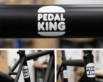 May include: Black bicycle frame with white "PEDAL KING" logo in a burger-like design. The logo is repeated on the frame and front post. The bicycle is black with a focus on the frame and branding.