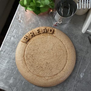 May include: A round, wooden bread board with the word "BREAD" carved into the top. The board has a circular groove and visible wear marks. Other kitchen items are in the background, including a small metal strainer and a copper pot with greenery.