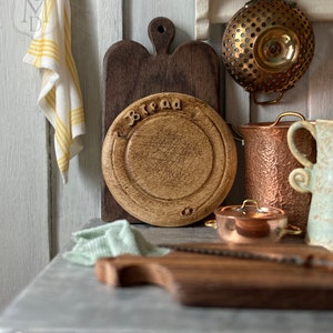 May include: A miniature kitchen scene with a wooden breadboard, a dark brown cutting board, and a copper pot. A yellow and white striped towel hangs on the left. The breadboard has the word "bread" carved into it.