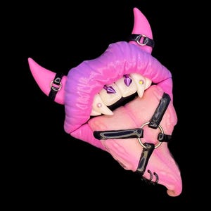 Puede incluir: Una boca esculpida de color rosa y morado con dientes afilados y una lengua, acentuada con gemas moradas y correas de cuero negro.