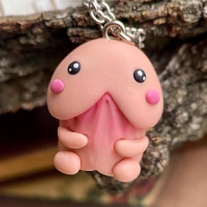 Könnte beinhalten: Eine skurrile Anhänger-Halskette mit einer rosa, cartoonartigen Figur. Die Figur hat schwarze Augen und rosa Wangen, mit einem hellrosa Kopf und einem dunkleren rosa Körper. Sie hängt an einer silberfarbenen Kette.