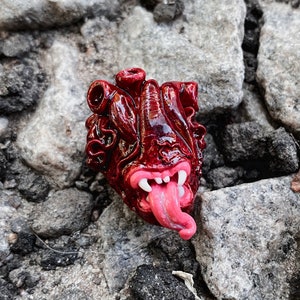 Peut inclure: Un cœur en céramique rouge détaillé avec une bouche ouverte et des dents, et une langue rose qui sort. Le cœur est texturé et a une finition brillante.