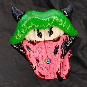 May include: A sculpted, stylized mouth with green lips, white teeth, and a pink tongue. Black horns and accents frame the mouth. The tongue has a piercing and a blood-like effect.