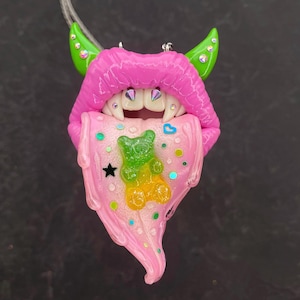 May include: A pink, dripping tongue with fangs, green horns, and a gummy bear inside. The tongue is covered in glitter and has a black star and a pink heart.