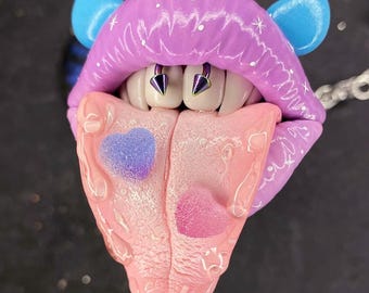 Könnte beinhalten: Ein lila und blauer Mund mit einer herausgestreckten rosa Zunge. Der Mund hat zwei silberne Nieten und die Zunge hat zwei rosa und blaue Herzen.