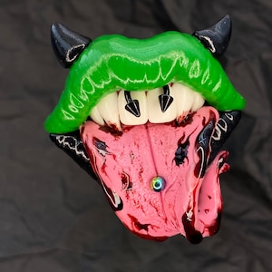 May include: A green and black clay sculpture of a mouth with fangs and a tongue. The tongue is pink and has a small, iridescent blue ring piercing.