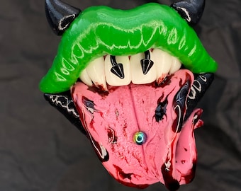 Könnte beinhalten: Eine grüne und schwarze Tonskulptur eines Mundes mit Zähnen und einer Zunge. Die Zunge ist rosa und hat ein kleines, schillerndes blaues Ringpiercing.