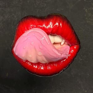 May include: Close-up of a pair of red lips with a pink tongue sticking out. The lips are glossy and the tongue is wet.