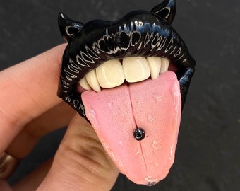 Könnte beinhalten: Schwarze, glänzende Skulptur eines Mundes mit weißen Zähnen und einer rosa Zunge. Die Zunge hat ein schwarzes Piercing. Die Lippen haben kleine Hörner an den oberen Ecken.