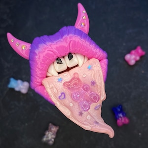 May include: A whimsical, fantasy-themed sculpture featuring a pair of pink and purple lips with fangs and horns. The tongue is a light pink color with a gummy bear design. Several gummy bear candies in various colors are scattered around the sculpture.