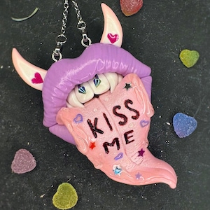 Puede incluir: Un colgante único con un diseño de labios morados, dientes blancos y una lengua rosa con la inscripción "KISS ME". El colgante tiene cuernos rosas con detalles de corazón y cuelga de una cadena plateada. Caramelos coloridos rodean el colgante.