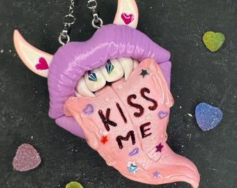 Könnte beinhalten: Ein einzigartiger Anhänger mit einem lila Lippen-Design, weißen Zähnen und einer rosa Zunge mit der Aufschrift "KISS ME". Der Anhänger hat rosa Hörner mit Herzakzenten und hängt an einer silbernen Kette. Bunte Süßigkeiten umgeben den Anhänger.