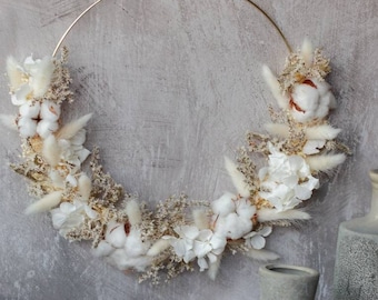 May include: A gold metal hoop wreath decorated with dried white cotton bolls, dried white flowers, and dried beige grasses.
